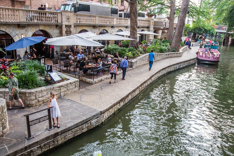 Umbrella-shaded patio tables beside the San Antonio River Walk with a river barge passing by